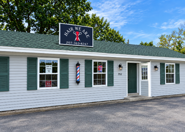 Gray clapboard storefront with the Hair…We Are! sign on the roof and a barber pole by the windows